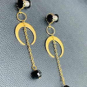 May include: A pair of black and gold earrings. Each earring features a black, round stud with a gold-colored decorative edge. A gold chain connects the stud to a gold crescent moon shape, with a dangling black bead at the end.