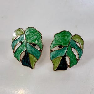 May include: A pair of gold-toned stud earrings in the shape of a monstera leaf. The leaves are divided into sections of varying shades of green, with a dark green border. The earrings have a glossy finish and a post backing.