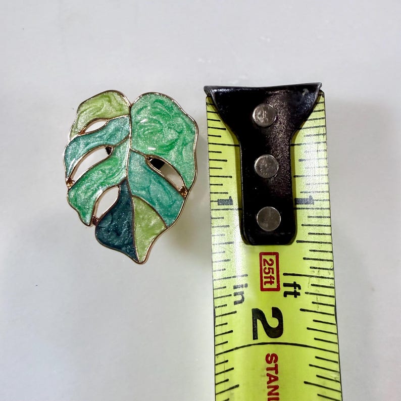 May include: A green and gold-toned leaf-shaped brooch. The brooch is a mix of light and dark green enamel with gold-colored trim. The brooch is approximately 2 inches in length.