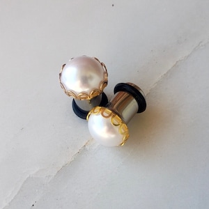 May include: Two silver ear gauges with a white pearl and gold trim. The gauges are on a white surface.