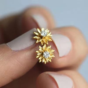 May include: A pair of gold-colored stud earrings, each shaped like a sunflower with delicate petals. The center of each flower features a sparkling, clear gemstone. The earrings are held between fingers against a soft background.