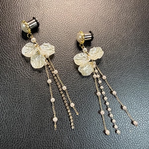 Può includere: Un paio di dilatatori pendenti con tunnel argentati sormontati da perle artificiali bianche. Ogni dilatatore presenta tre petali di fiore in acrilico bianco e diverse catenelle dorate decorate con piccole perle bianche e cristalli trasparenti.