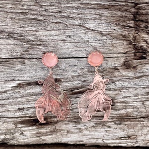 May include: A pair of rose gold earrings featuring a delicate, cutout design of a betta fish with a pink stone accent.