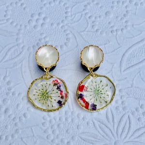 May include: A pair of dangle earrings with a gold-colored trim. The earrings feature a clear resin with dried flowers in shades of red, purple, and white. The top portion is a white, pearlescent stud.