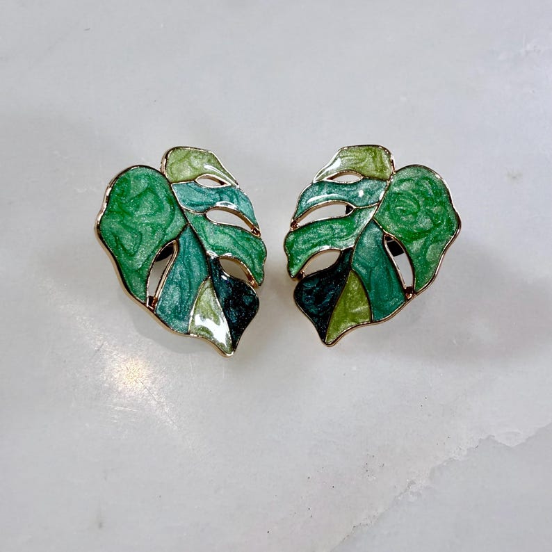May include: A pair of gold-toned stud earrings in the shape of Monstera leaves. The leaves are enameled in shades of green, from light to dark, with a glossy finish. The earrings have a botanical theme.