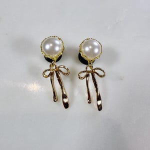 May include: A pair of gold-toned earrings featuring a large, round, faux pearl at the top, surrounded by a gold filigree. Below the pearl is a gold bow design with dangling ribbons. The earrings are on a white surface.