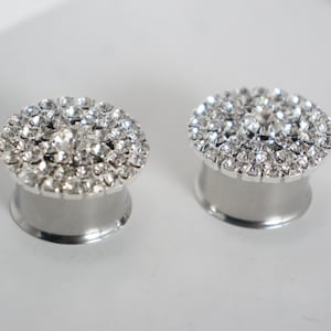 May include: A pair of silver ear gauges with a circular design featuring a large cluster of clear rhinestones.