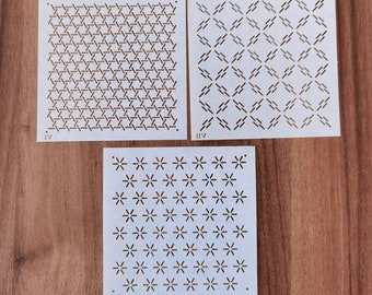 May include: Three white stencils with geometric patterns, including a hexagonal design, a diamond pattern, and a floral arrangement. The stencils are labeled with Roman numerals and are displayed on a wooden surface.