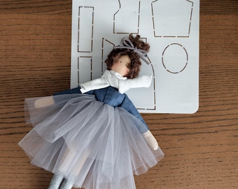May include: A handmade doll with a blue dress and a gray tulle skirt, posed next to a white stencil with clothing pattern outlines. The doll has brown hair and a gray bow.