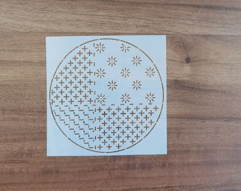 May include: A square, off-white stencil featuring a circular design with various patterns. The design includes cross-stitch and starburst shapes, cut out to create the pattern. The stencil is made of a thin, flexible material.