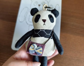 May include: A handmade panda bear keychain with black and white fabric, black arms and legs, and a small purse. The panda has black eyes, ears, and a scarf. The purse is purple with a blue and yellow bow.