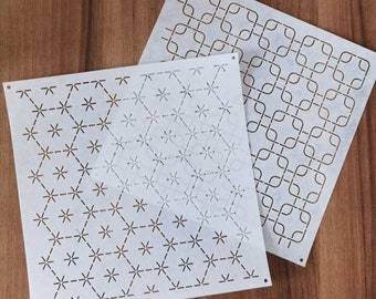 May include: Two white stencils with geometric patterns. One features a repeating hexagonal design with star-like shapes, and the other has a square and curved line pattern. Both stencils have small holes in the corners.