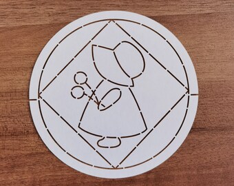 May include: A white paper stencil featuring a stylized figure of a person in a bonnet holding flowers. The design is within a circle and a diamond shape, with cut-out lines. The stencil is on a wooden surface.