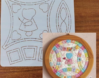 May include: A quilting stencil with a fairy design and a finished quilt in a wooden frame. The stencil is light blue with cut-out shapes of a fairy, hearts, and geometric patterns. The quilt features a fairy design and colorful fabric patches.