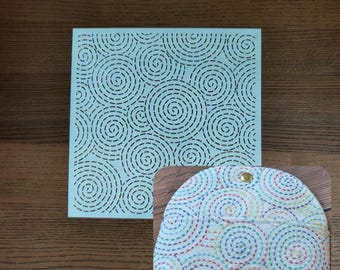 May include: A light blue square with a repeating spiral design and a small, rounded pouch. The pouch is white with a colorful spiral pattern and a gold snap closure. The items are on a wooden surface.