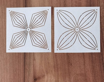 May include: Two white stencils with a floral design. One stencil features a starburst pattern, while the other has a four-petal flower design. The stencils are placed on a wooden surface.