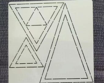 May include: A white stencil with a geometric design of triangles. The stencil features several triangles of varying sizes, outlined with dashed lines. The design is centered on a square, creating a pattern for crafts or art projects.