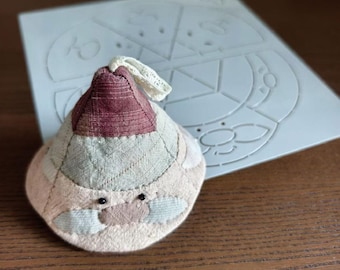 May include: A handmade fabric Santa Claus ornament with a burgundy hat and a cream-colored beard. The ornament is made of various fabric patches in shades of pink, beige, and gray. A white lace loop is attached to the top. A white template is in the background.