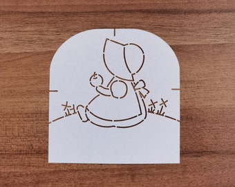 May include: A white stencil featuring a seated figure in a bonnet holding a small fruit. The figure is wearing a dress with a bow. The stencil has cut-out details and includes text in a foreign language.