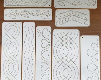 May include: A collection of white quilting stencils with various designs, including floral patterns, hearts, and geometric shapes. The stencils are rectangular and feature cut-out designs for creating decorative stitching on fabric. The stencils are arranged on a wooden surface.