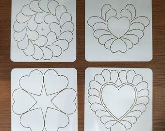 May include: Four white quilting stencils with different heart and leaf designs. The stencils are square and feature cut-out patterns for creating decorative quilt patterns. The designs include hearts, leaves, and a star.