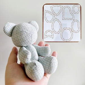 May include: A handmade gray teddy bear with a plaid pattern is held in a hand. Next to the bear is a wooden template with cut-out shapes for sewing the bear. The bear is made of soft fabric and has small ears.