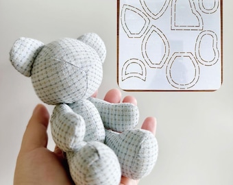 May include: A handmade gray teddy bear with a plaid pattern is held in a hand. Next to the bear is a wooden template with cut-out shapes for sewing the bear. The bear is made of soft fabric and has small ears.