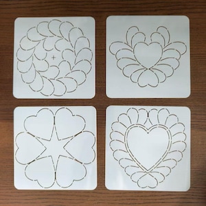 May include: Four white quilting stencils with various heart and leaf designs. The stencils are square and feature cut-out patterns for creating decorative quilt patterns. Designs include a heart with a leaf border and a wreath of leaves.
