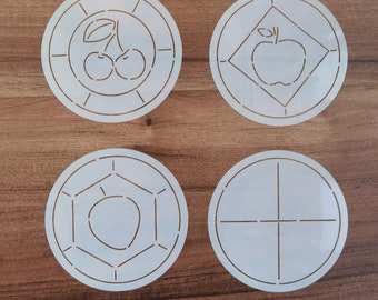 May include: Four white circular stencils with fruit and geometric designs. One stencil features cherries, another an apple, a third an abstract shape, and the last a cross design. The stencils are on a wooden surface.
