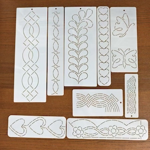 May include: A collection of white plastic stencils with various decorative designs, including geometric patterns, hearts, butterflies, and floral motifs. The stencils are arranged on a wooden surface, offering a variety of creative options for crafts and art projects.