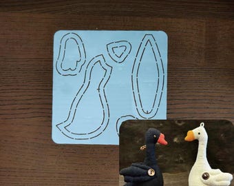May include: A blue cutting template with dashed lines for a goose-shaped plushie, alongside two finished plush geese. One goose is black with a red beak and feet, the other is white with orange accents.