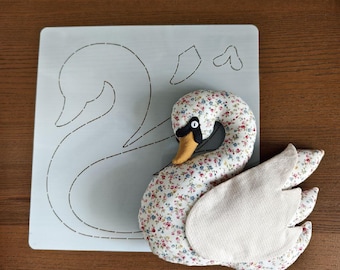 May include: A handmade swan plush toy with a floral patterned body, a yellow beak, and white wings. The swan is next to a light blue template with a swan outline. The background is a wooden surface.