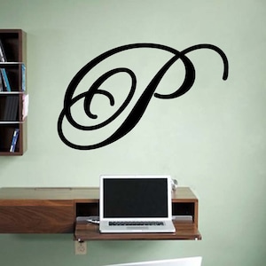 May include: A large, black, cursive letter "P" is affixed to a light green wall. The letter is the focal point, with a laptop on a wooden desk below. A bookshelf is visible to the left.