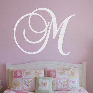 May include: White vinyl wall decal of a cursive letter M with a decorative swirl. The decal is on a light pink wall above a white bed with a floral patterned quilt.