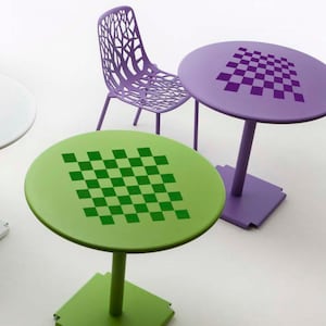 May include: Two round tables with a checkerboard pattern on top. One table is green with a green checkerboard and the other is purple with a purple checkerboard. A purple chair with a unique design is in the background.