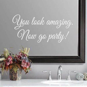 May include: A mirror with a black frame displays the text "You look amazing. Now go party!" in a white, cursive font. A floral arrangement in a decorative vase sits below the mirror, with a sink and faucet visible.