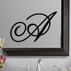 May include: A black, cursive letter "A" displayed on a mirror. The letter features a stylized, elegant design with a swirling loop and a diagonal stroke. The mirror has a dark frame, and the background includes a bathroom sink and floral arrangement.