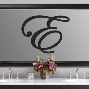 May include: A large black framed mirror with a gray surface, featuring a black cursive letter "E" in the center. The mirror is above a white countertop with two sinks, faucets, and a floral arrangement.