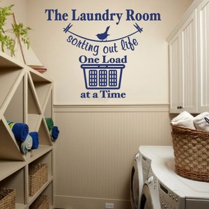 May include: A white laundry room with a blue vinyl wall decal that reads "The Laundry Room #sorting out life One Load at a Time". The decal features a bird on a clothesline with clothespins. There are two white washing machines and a wicker basket with towels.