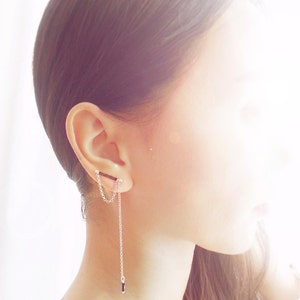 May include: Silver ear cuff with a dangling chain and a small black bead. The ear cuff is attached to a chain that hangs down. The jewellery is worn on the ear.