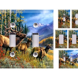 May include: A set of light switch covers featuring a scenic image of a herd of elk in a forest setting. The elk are brown and have large antlers. The background is a colorful autumn forest with mountains in the distance.