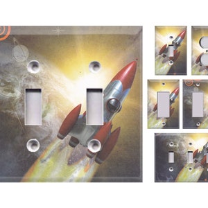 May include: A set of light switch covers featuring a retro-style rocket ship flying through space. The rocket is red and white with a yellow flame trail. The background is a dark blue with stars and planets.