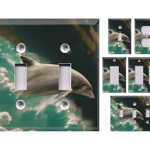 May include: Decorative light switch covers featuring a dolphin swimming through a cloudy sky. The main cover has two light switches and three decorative bubbles. Additional covers include single and double light switches and an electrical outlet.