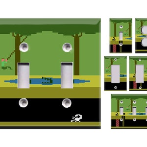 May include: A set of light switch covers featuring a pixelated video game design. The design includes a green jungle scene with a character swinging on a vine, a crocodile in a river, and a character running on the ground. The switch covers are black with white outlines.