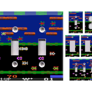 May include: A set of light switch covers featuring a retro video game design. The covers are blue, green, and black with pixelated graphics of a car, a turtle, and a frog. The text "1-UP" is visible on the bottom of the cover.