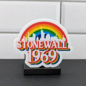 May include: A white sticker with a rainbow and the text "Stonewall 1969" on a black base. The sticker features a silhouette of the New York City skyline.