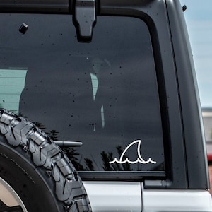 May include: A white shark fin decal on a black car window. The decal is a simple line drawing of a shark fin with a wave underneath.