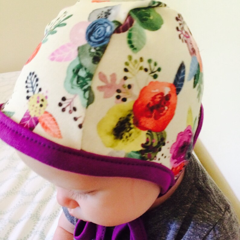 May include: A white baby bonnet with a floral print in shades of pink, red, yellow, green, and blue. The bonnet has a purple trim.
