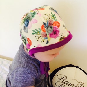 May include: A baby wearing a white bonnet with a floral print and a purple trim. The bonnet has a tie at the back.