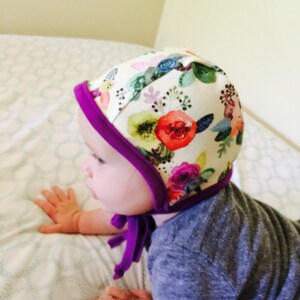 May include: A baby wearing a white and purple floral print bonnet with a purple tie. The bonnet has a white background with a watercolor floral pattern.